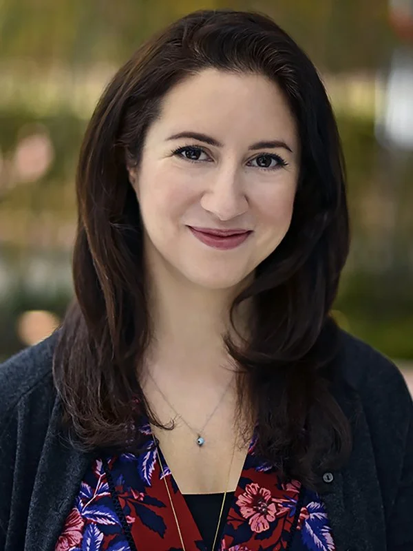 A woman with dark brown hair smiles warmly. She wears a floral blouse and a dark cardigan. The background is softly blurred, conveying a pleasant ambiance.