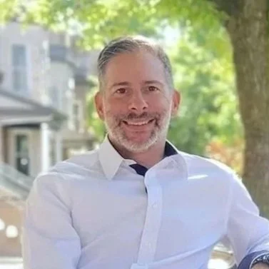 Smiling man with gray hair and beard in a white shirt sits outdoors with blurred leafy background, conveying a relaxed and cheerful vibe.