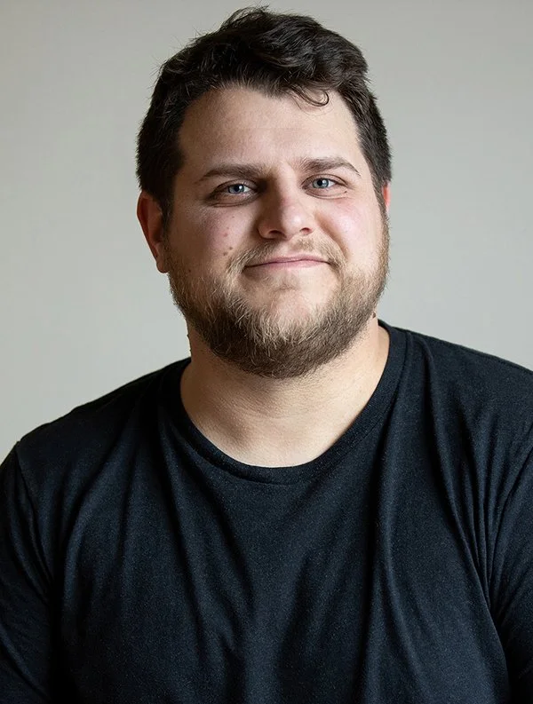 A man with short dark hair and a beard smiles gently, wearing a black T-shirt, in front of a neutral background. The mood is relaxed and approachable.