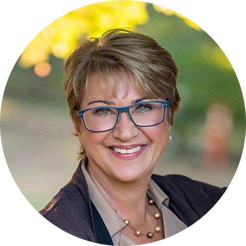 A woman with short hair and blue glasses smiles warmly. She wears a blazer, a blouse, and a beaded necklace. Blurred green and yellow background.