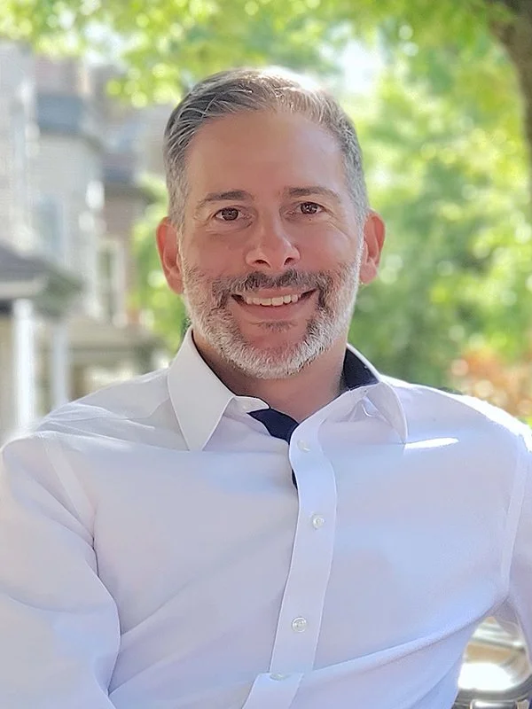 Smiling man with gray hair and beard in a white shirt sits outdoors with blurred leafy background, conveying a relaxed and cheerful vibe.