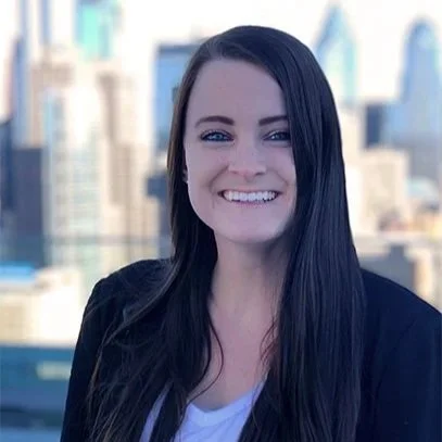 Smiling woman with long brown hair stands outdoors against a blurred urban skyline, wearing a black blazer and white shirt, conveying a cheerful mood.
