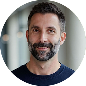 Man with a beard and short dark hair smiling warmly, wearing a navy sweater, standing in a bright hallway with soft lighting. Calm and friendly mood.