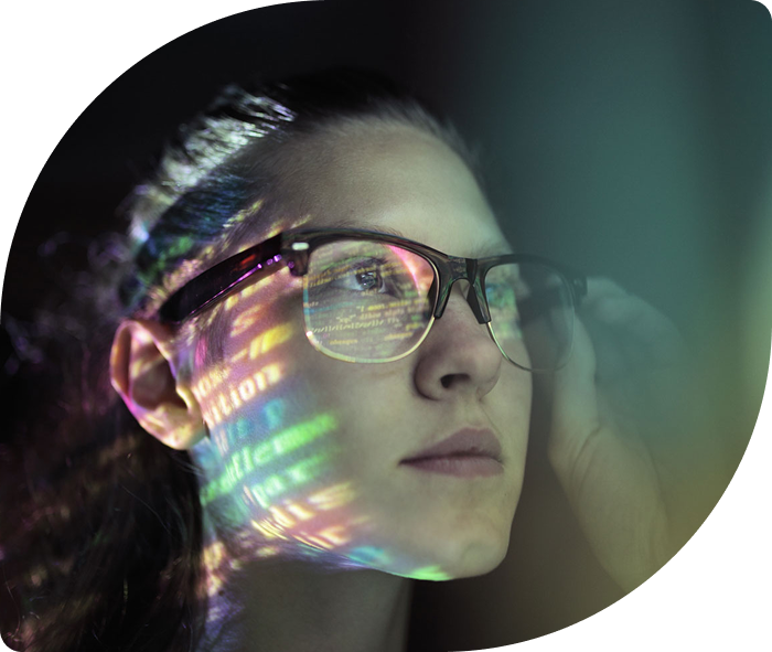 A person wearing glasses looks intently at a screen, with colorful computer code reflecting on their face. The scene conveys focus and engagement with technology.