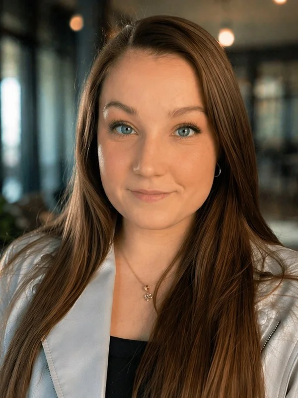 A young woman with long brown hair and blue eyes looks into the camera. She wears a light-colored jacket and a delicate necklace, with a calm expression. Blurred background.