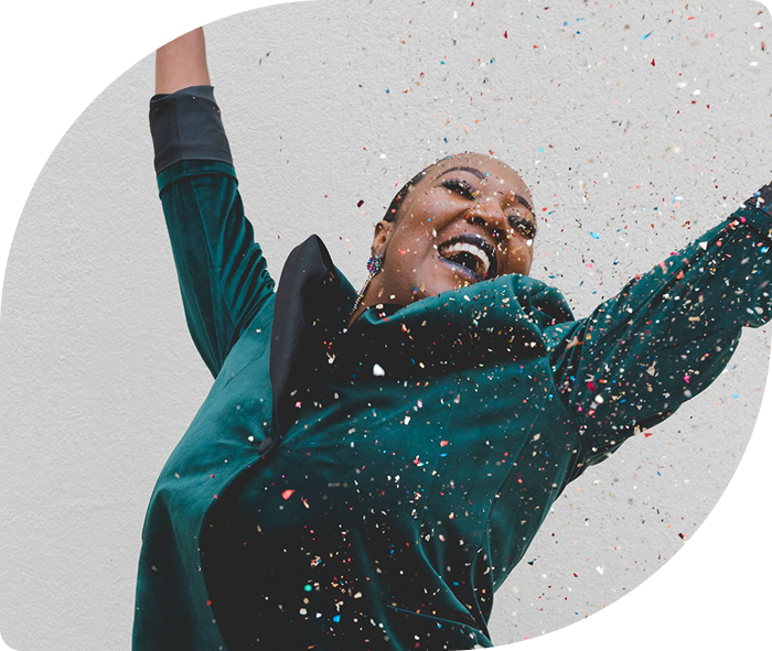 Woman joyfully celebrating, throwing colorful confetti into the air. She wears a green jacket and smiles widely against a plain background. Energetic and festive mood.