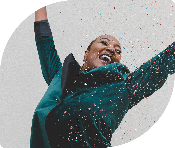Woman joyfully celebrating, throwing colorful confetti into the air. She wears a green jacket and smiles widely against a plain background. Energetic and festive mood.