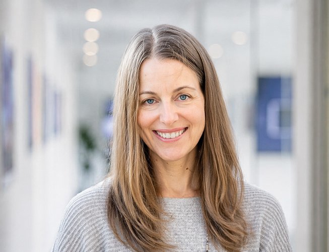 Smiling woman with long brown hair wearing a gray sweater. Soft, light-filled background with blurred lights, conveying warmth and friendliness.