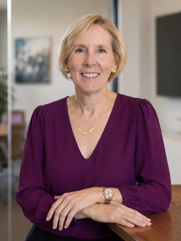 A smiling woman with short blonde hair wears a purple blouse and gold jewelry. She rests her hands on a wooden table, exuding a warm and professional demeanor.