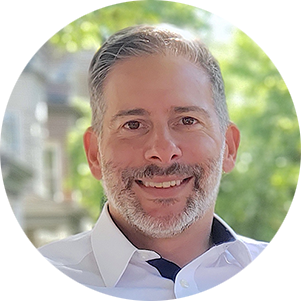 Smiling man with gray hair and beard in a white shirt sits outdoors with blurred leafy background, conveying a relaxed and cheerful vibe.