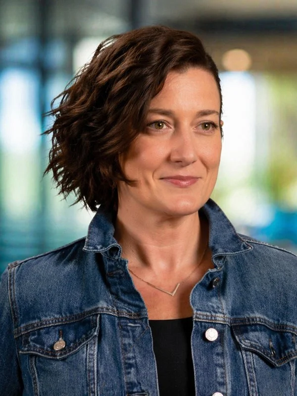 Smiling woman with short, wavy brown hair in a denim jacket, standing in a softly blurred indoor setting, exuding a confident and warm demeanor.