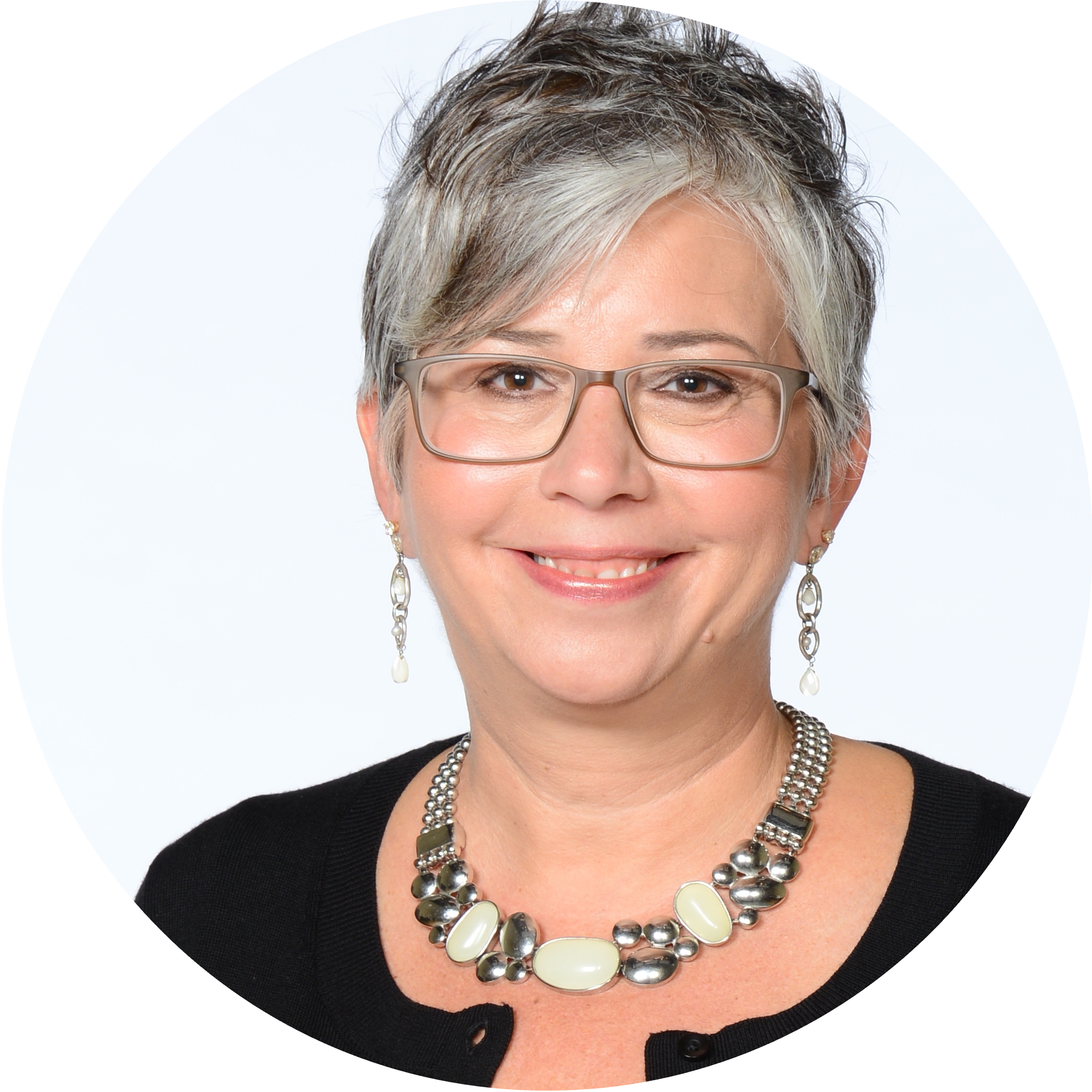 A woman with short gray hair smiles warmly at the camera. She wears glasses, a black top, dangling earrings, and a chunky beaded necklace, exuding confidence.