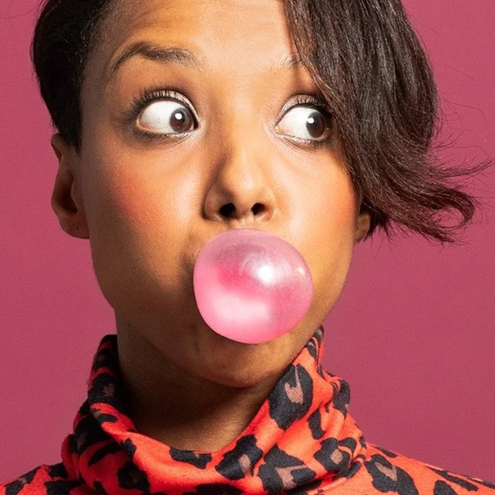 A woman with wide eyes blows a pink bubblegum bubble. She wears a red patterned sweater and stands against a matching pink background, conveying surprise.