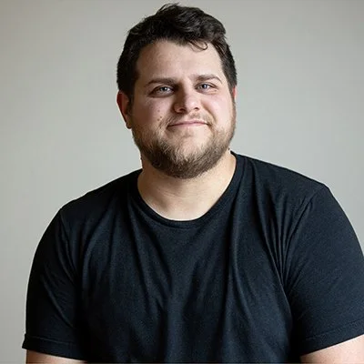 A man with short dark hair and a beard smiles gently, wearing a black T-shirt, in front of a neutral background. The mood is relaxed and approachable.