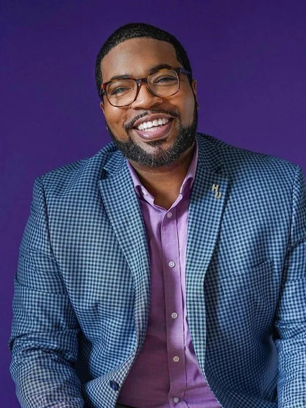Smiling man in glasses and a purple shirt with a checked blazer against a purple background, conveying a friendly and approachable tone.