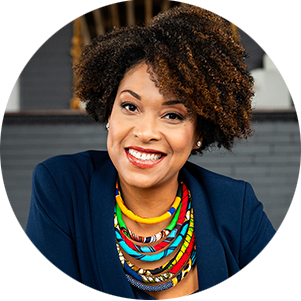 Smiling woman with curly hair wearing a navy blazer, colorful beaded necklaces, and rings, sitting on a brown sofa. The atmosphere is warm and inviting.