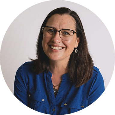 Smiling woman with glasses, wearing a blue shirt and earrings, against a plain white background. The tone is cheerful and welcoming.
