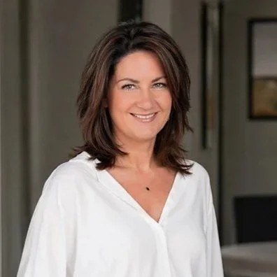 Woman smiling in a bright room, wearing a white blouse and dark pants. Her hands are in her pockets, conveying confidence and warmth.
