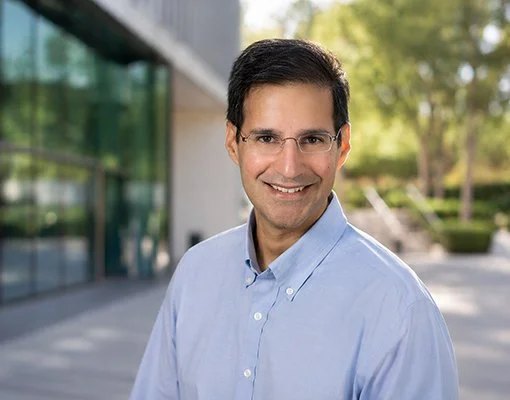 Smiling person wearing glasses and a light blue shirt stands outdoors with greenery and building in the background, conveying a friendly tone.