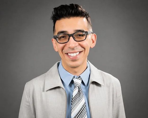 Smiling man with glasses and short dark hair, wearing a light gray blazer, blue shirt, and patterned tie. Gray background. Professional and cheerful tone.