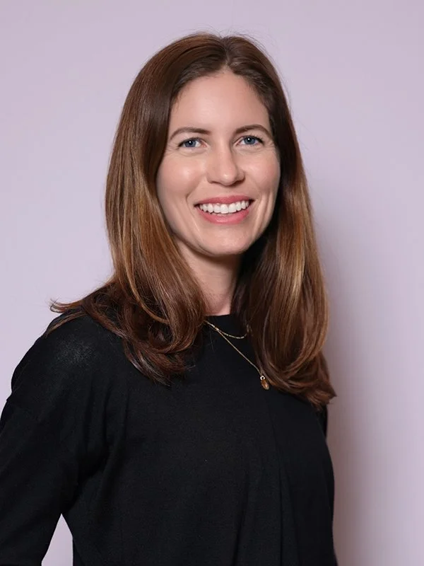 A smiling woman with long brown hair wearing a black top and necklace poses in front of a plain light purple background, conveying a friendly and approachable tone.