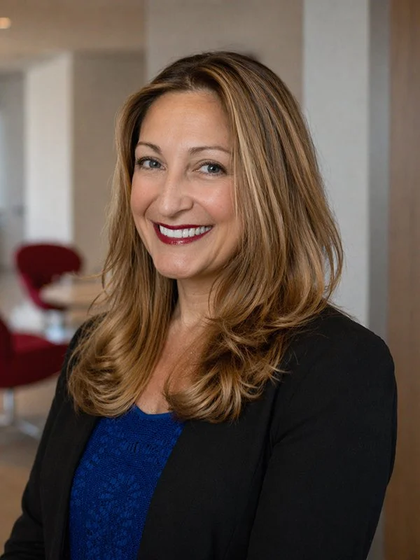 A woman with long brown hair and red lipstick smiles warmly, wearing a blue top and black blazer. The background is a softly-lit, modern office space.