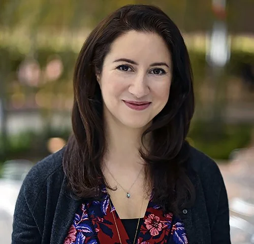 A woman with dark brown hair smiles warmly. She wears a floral blouse and a dark cardigan. The background is softly blurred, conveying a pleasant ambiance.