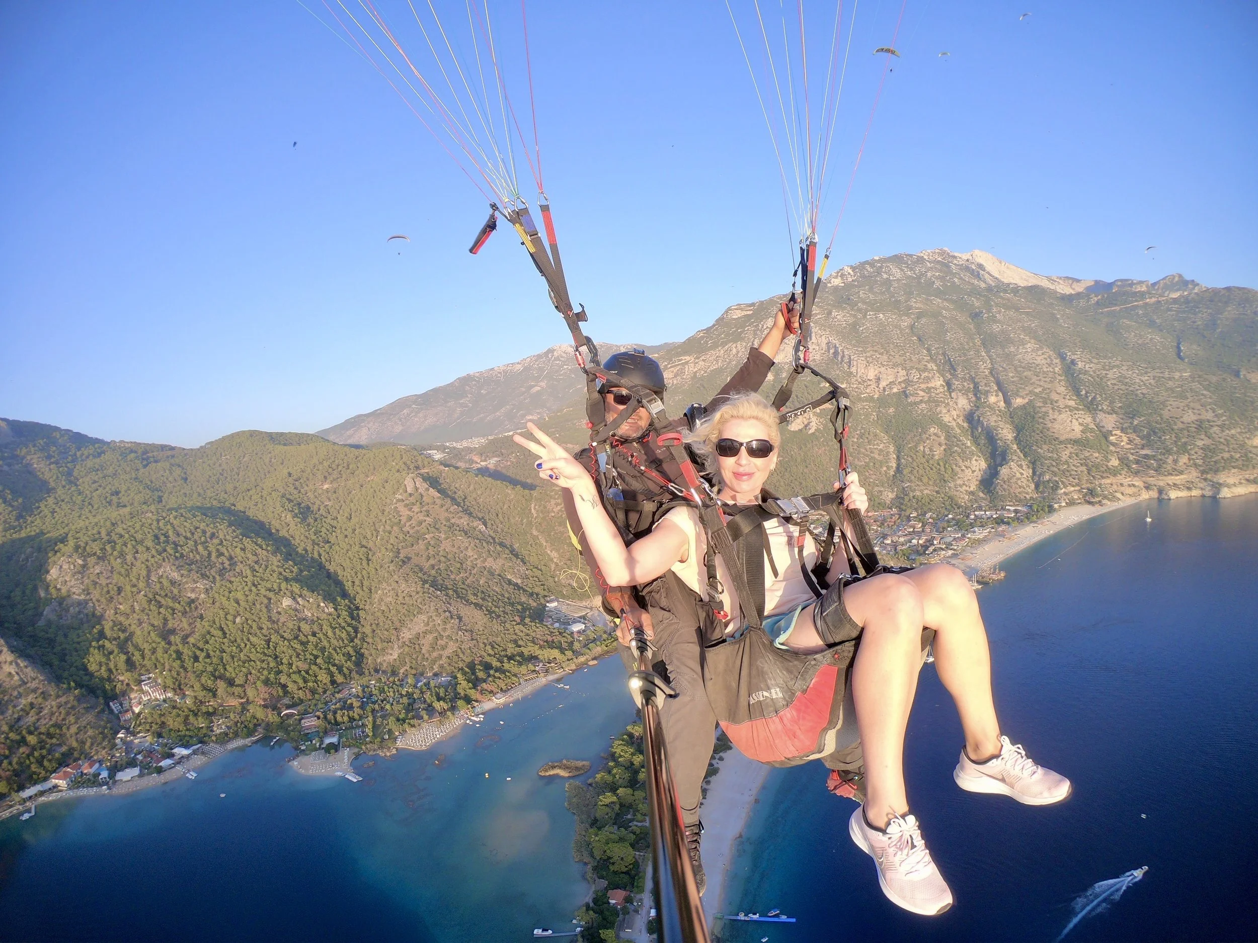 Two people paragliding over a scenic landscape with mountains, forests, a river, and a town below, during daytime with clear blue sky.