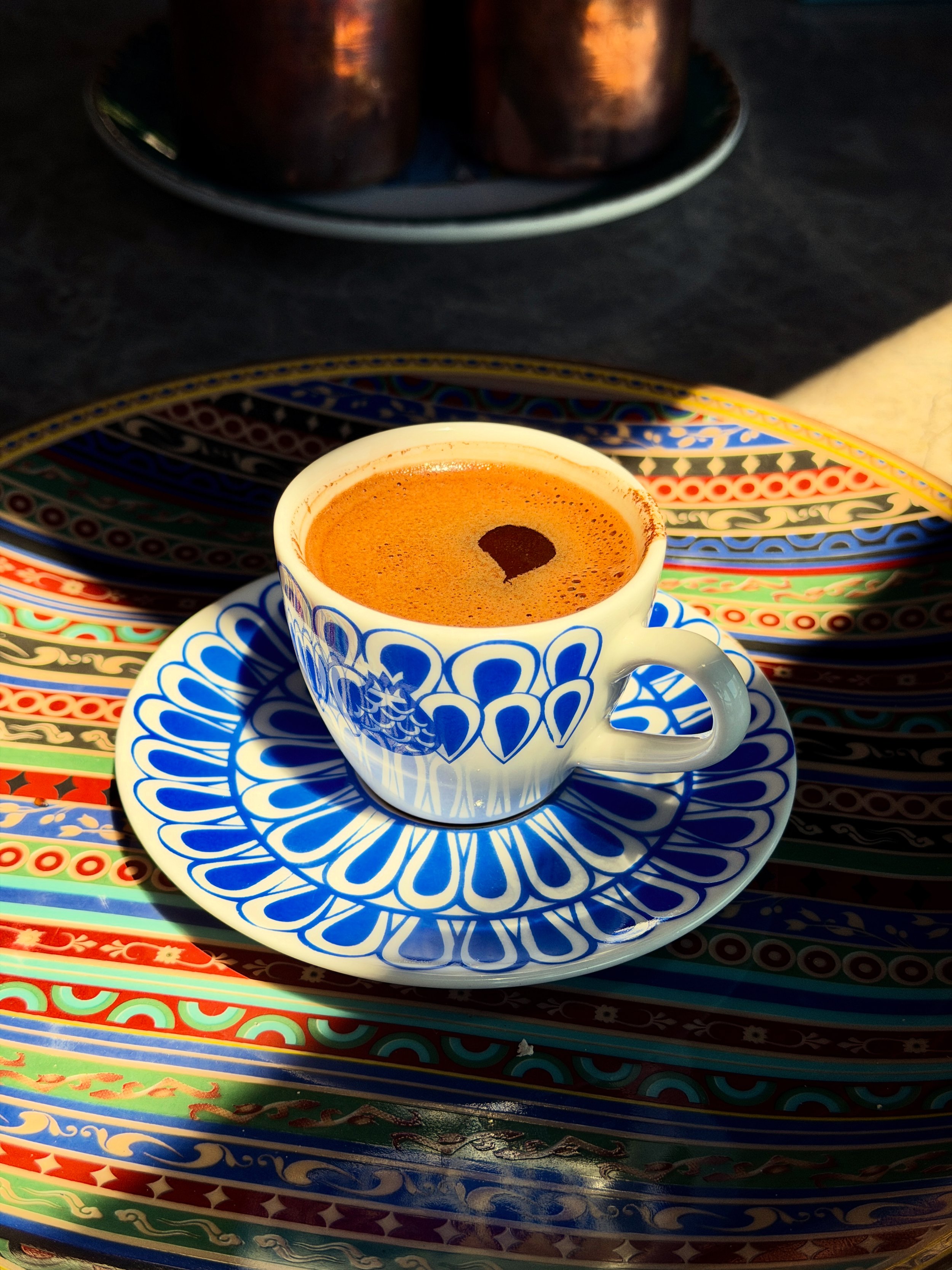 A cup of Turkish coffee with foam on top, placed on a patterned saucer, with a colorful decorative tray underneath, and part of a tray with two glasses in the background.
