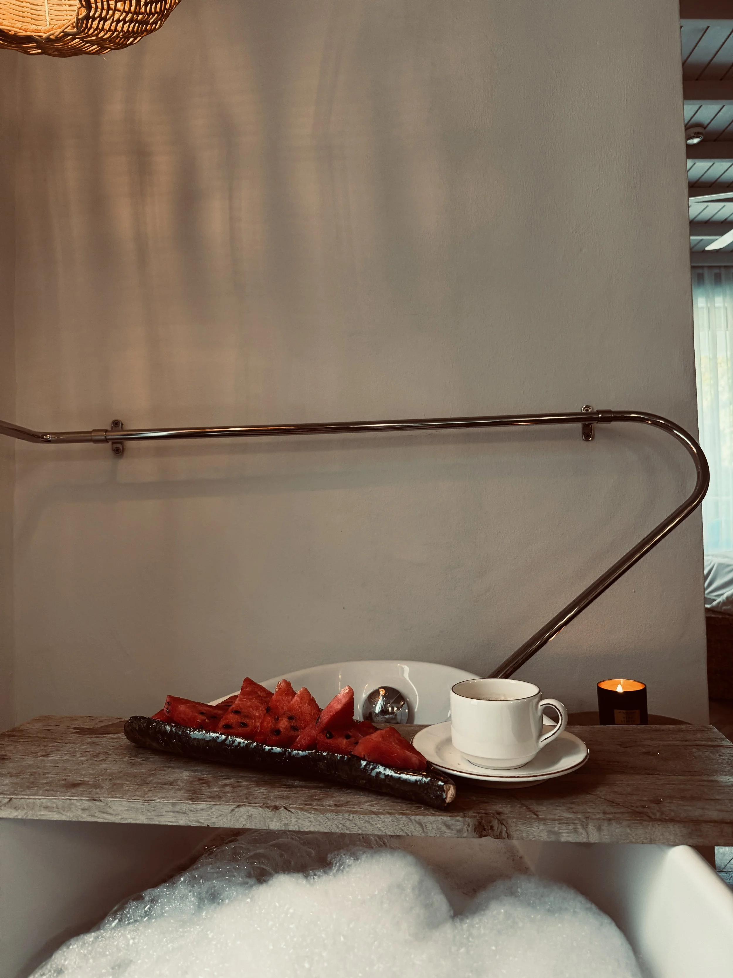 View of a table set for breakfast with watermelon slices on a black plate, a white cup and saucer, and a small orange candle. In the background, a bathtub filled with bubbly water is visible, with the edge of a wooden table and part of a wall.