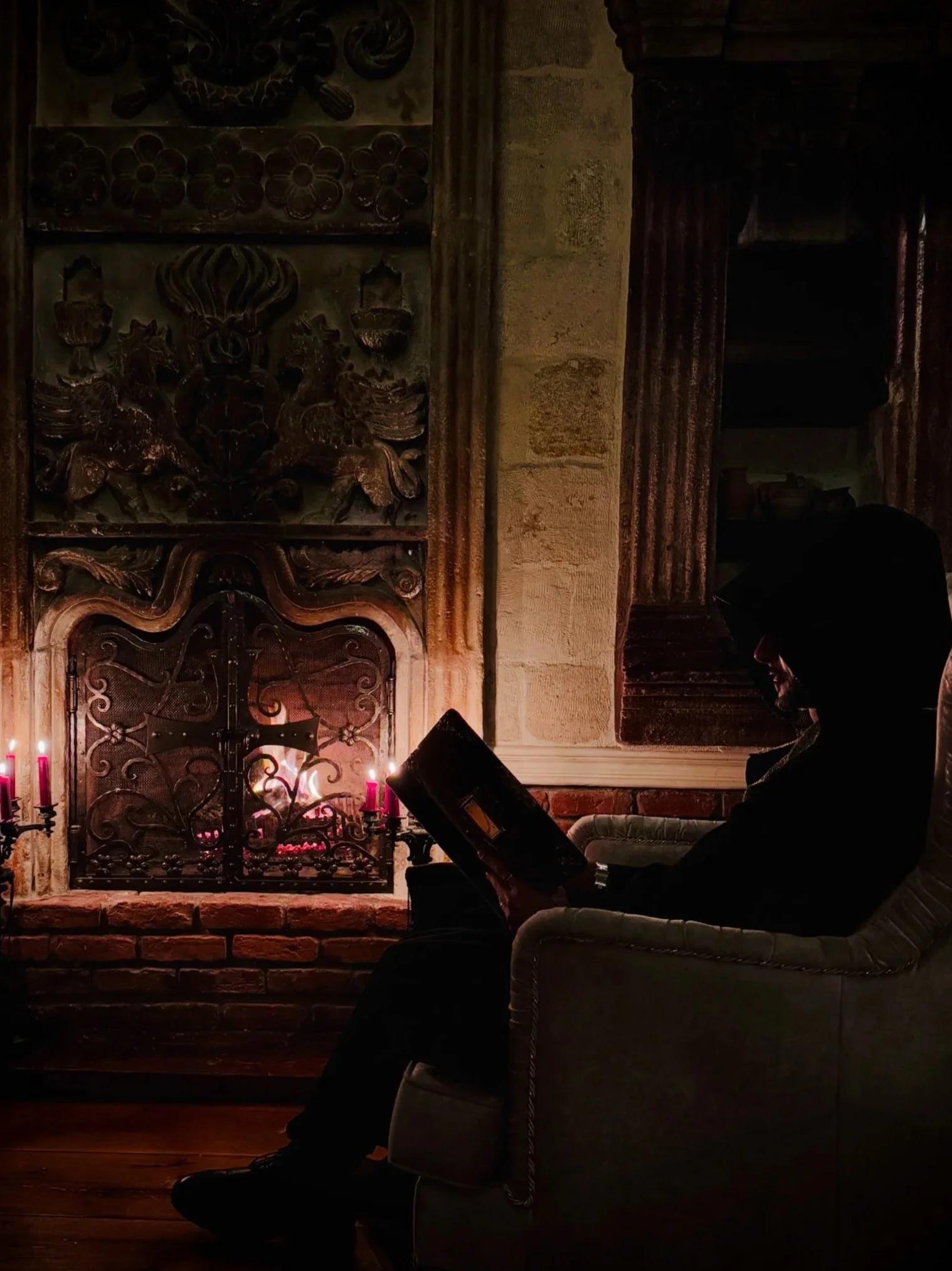 A person sitting in a chair by a fireplace, reading a book, with candles lit nearby in a dimly lit room.