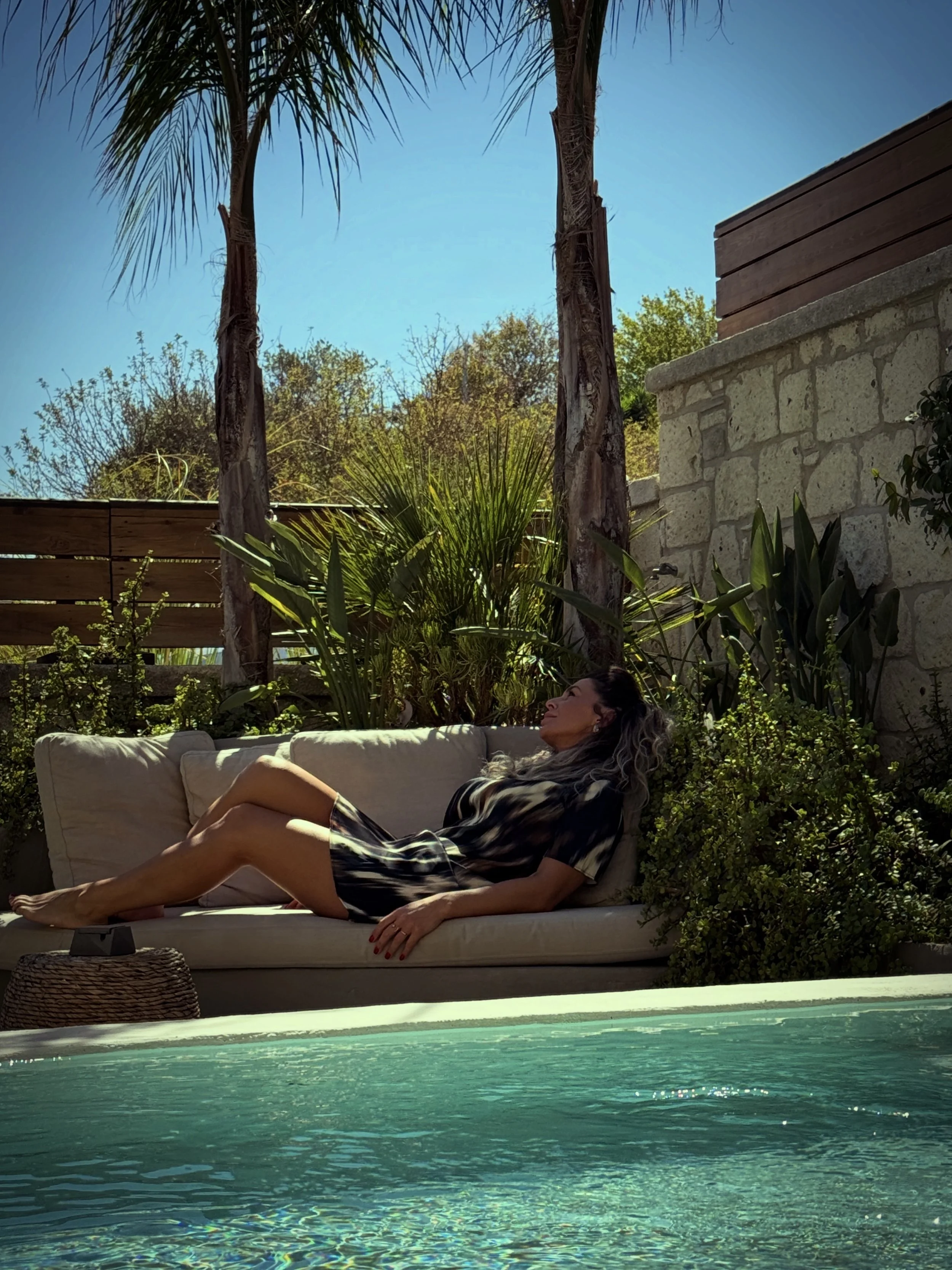 A woman relaxing on a beige outdoor sofa beside a swimming pool with clear water, lush green plants, trees, a wooden privacy fence, and a stone wall in a backyard on a sunny day.