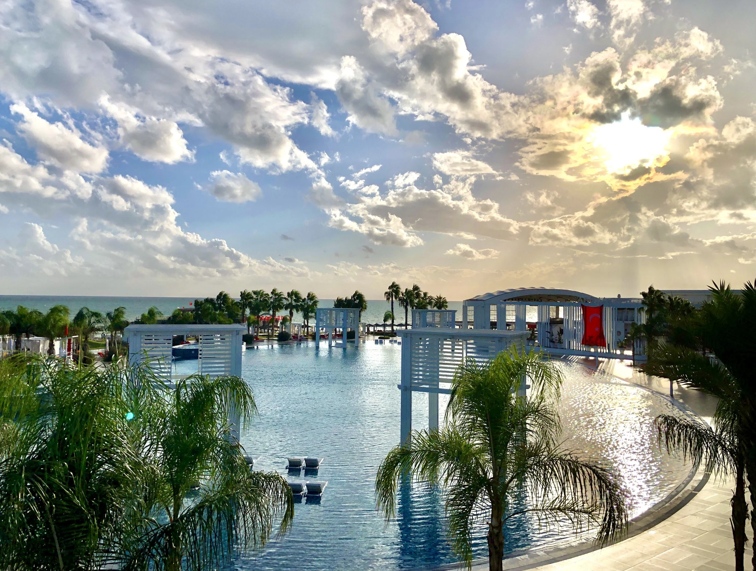 A poolside area near the ocean with lounge chairs, palm trees, and a swimming pool, under a partly cloudy sky with sunlight.