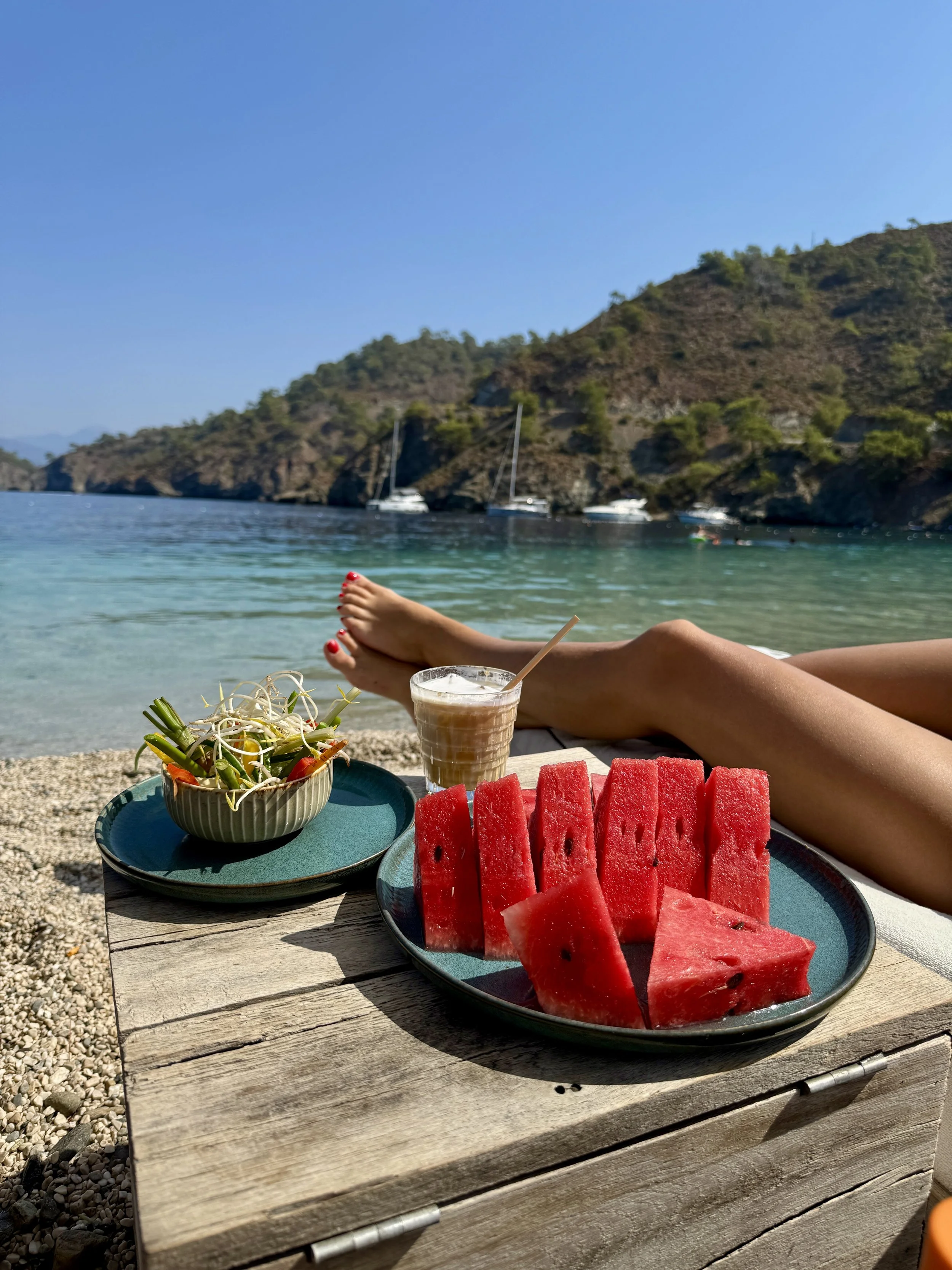 Scenic outdoor setting with a wooden table showcasing sliced watermelon, a tropical salad, and a smoothie, with a person relaxing by the water and sailboats at a hilly, green coastline under a clear blue sky.