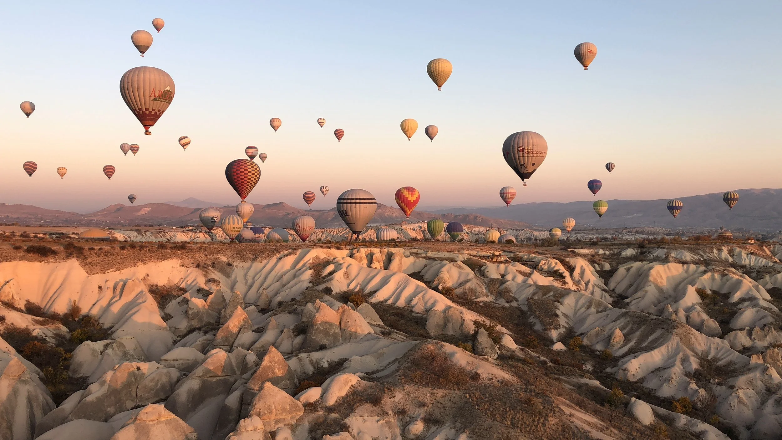 Multiple hot air balloons floating over a rocky, hilly landscape during sunrise or sunset.
