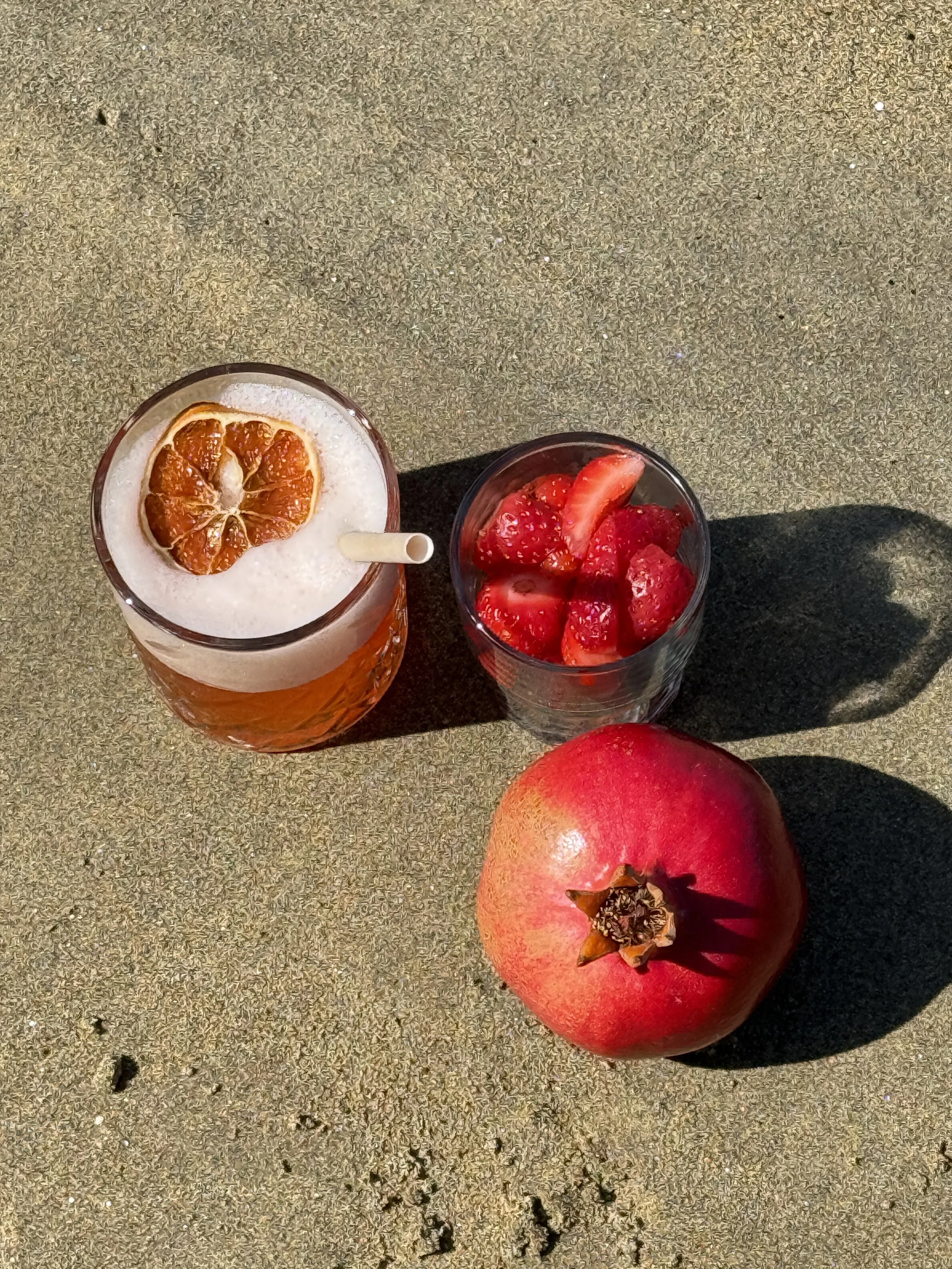 A pomegranate fruit, a glass of strawberry juice with ice and a slice of orange on top, and a glass of strawberries, all placed on a sandy surface.