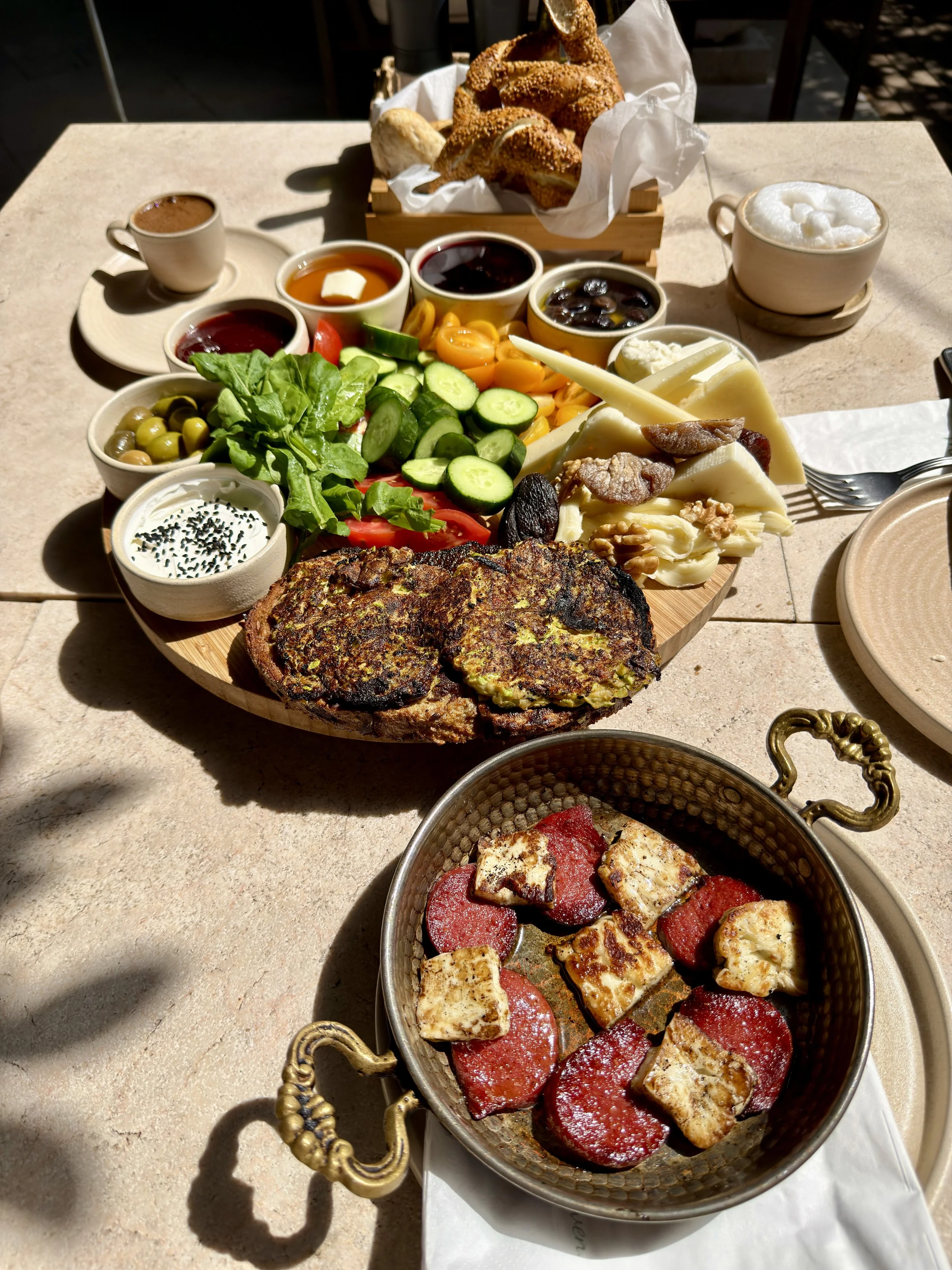 A breakfast spread on a table including a platter of sliced tomatoes, cucumbers, various cheeses, leafy greens, olives, and several small bowls of condiments or jams, alongside a basket of assorted bread and pastries, and cups of coffee or tea.