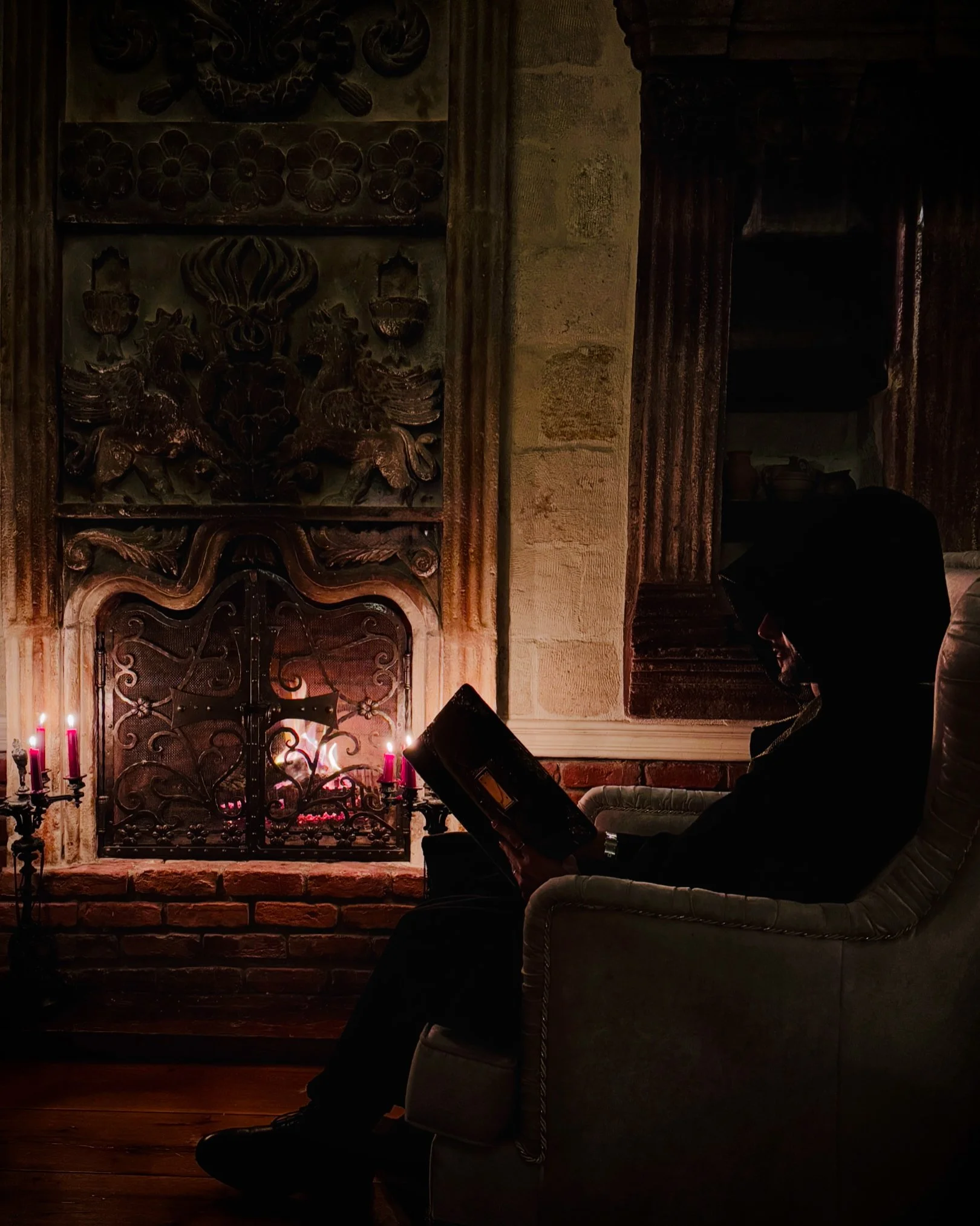 A person sitting in a chair reading a book by a lit fireplace with pink candles nearby in a dimly lit room.