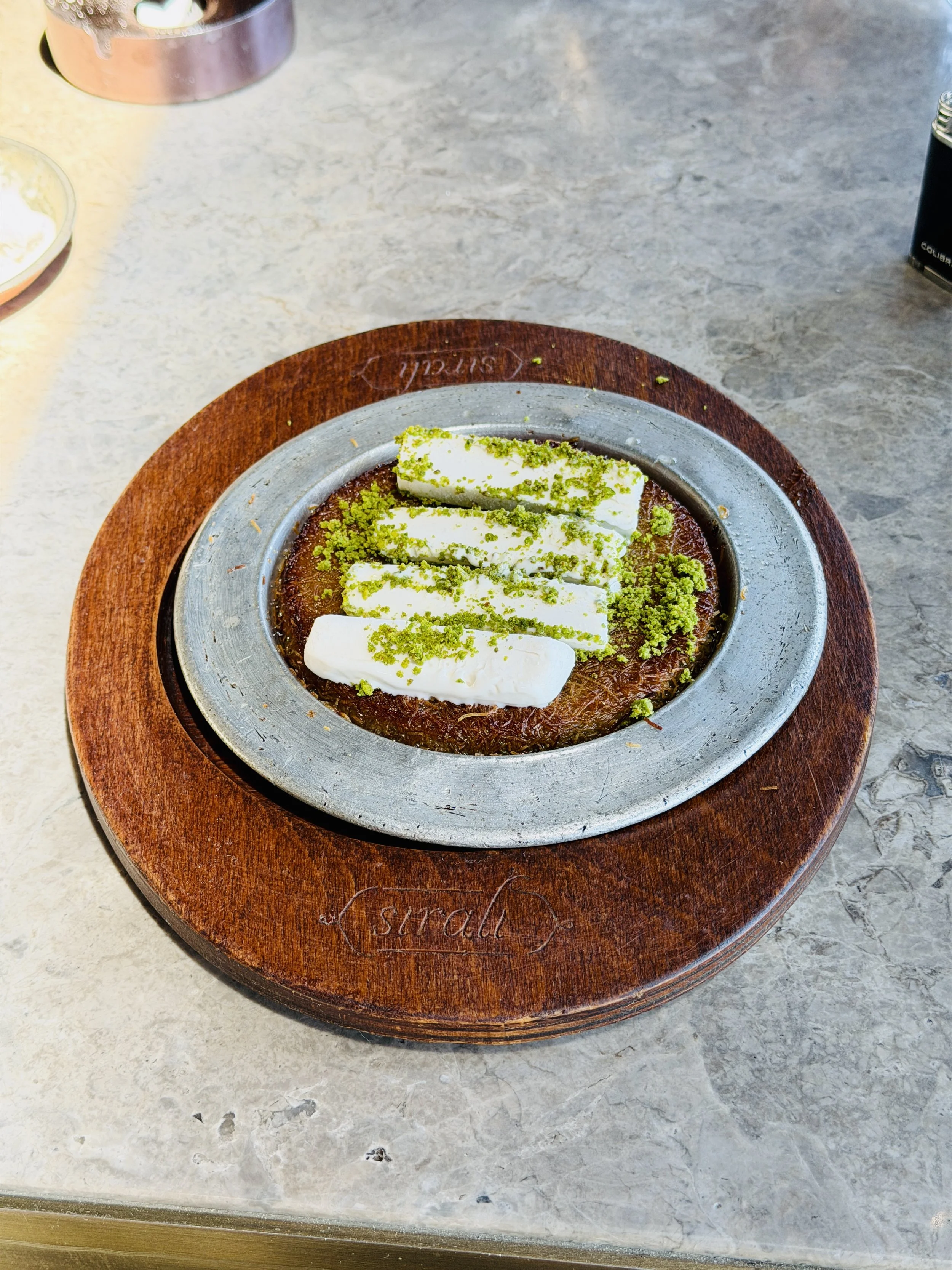 A dessert on a metallic plate with a layer of cream and green sprinkles, placed on a wooden round tray with the word 'sugar' engraved on it, on a gray marble surface.