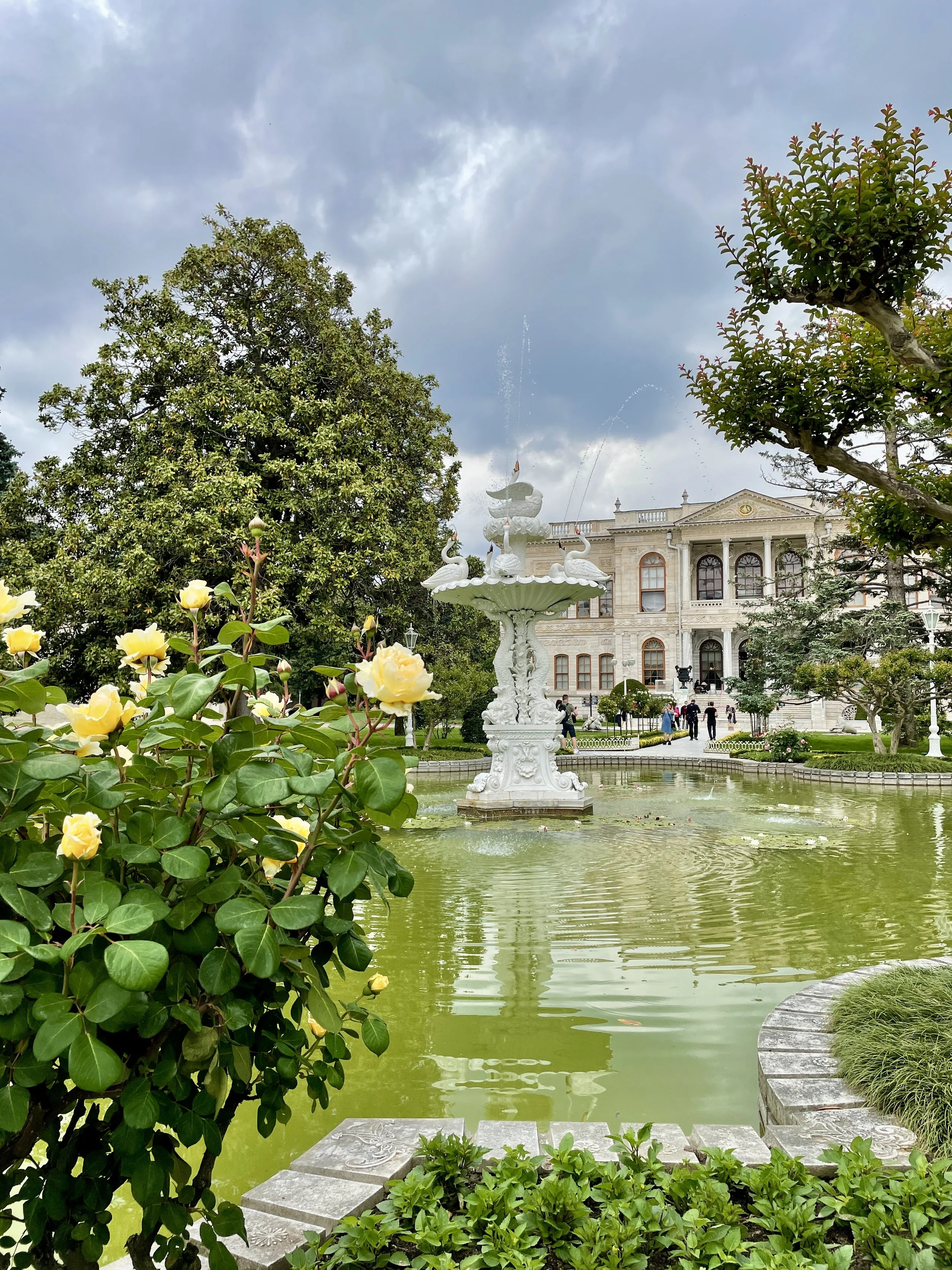 A decorative white fountain with swan-shaped figures in the center of a small pond, surrounded by greenery and a historic building in the background.