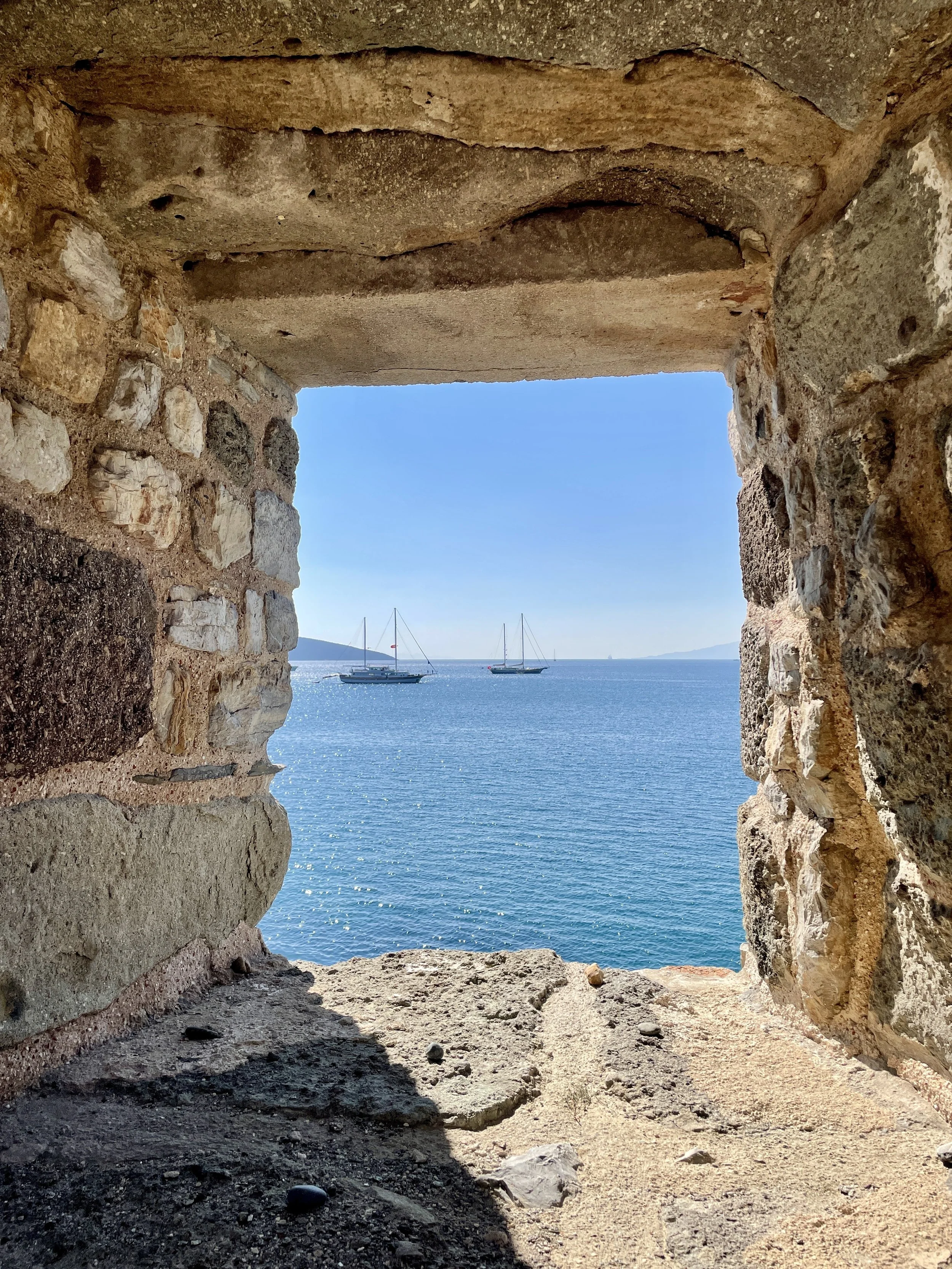 View of the ocean with sailboats through an opening in an old stone wall, under a clear blue sky.