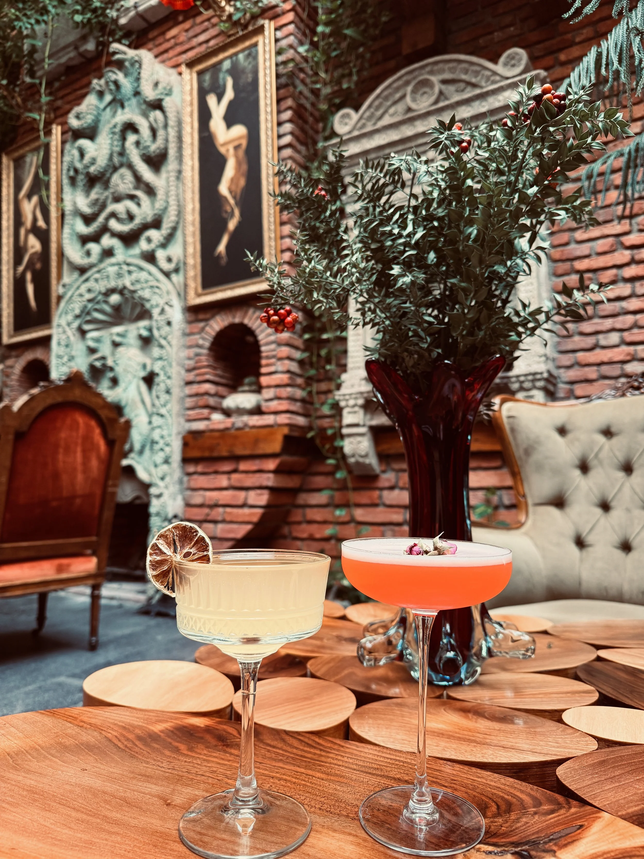 Two cocktails on a wooden table with decorative plates and a vase of greenery in a vintage-style room with brick walls and framed artwork.