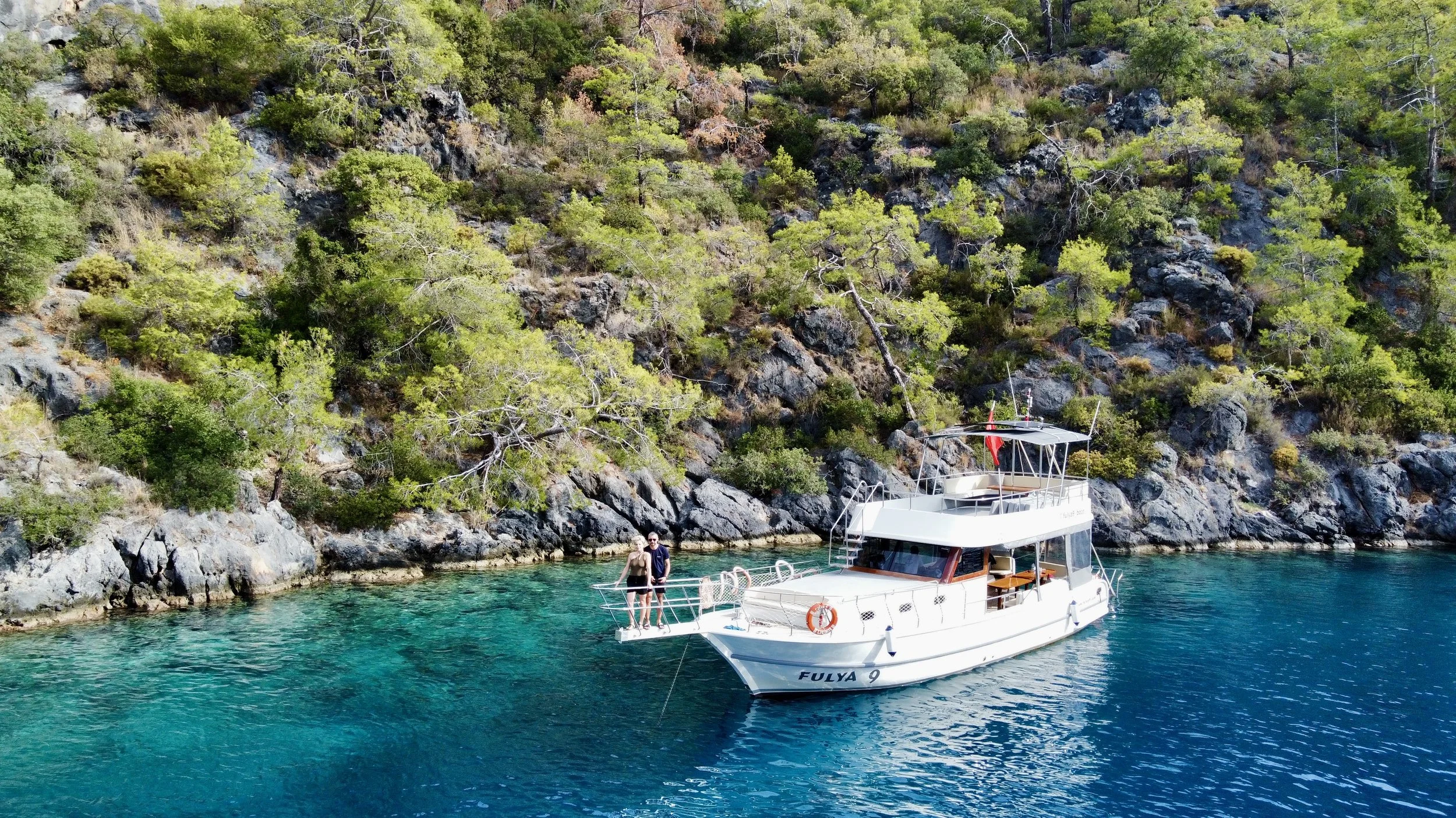 A white yacht named Fulya 9 is anchored in clear turquoise water near a rocky shoreline covered with green trees. Two people are standing on the yacht's deck, smiling and enjoying the sunny day.