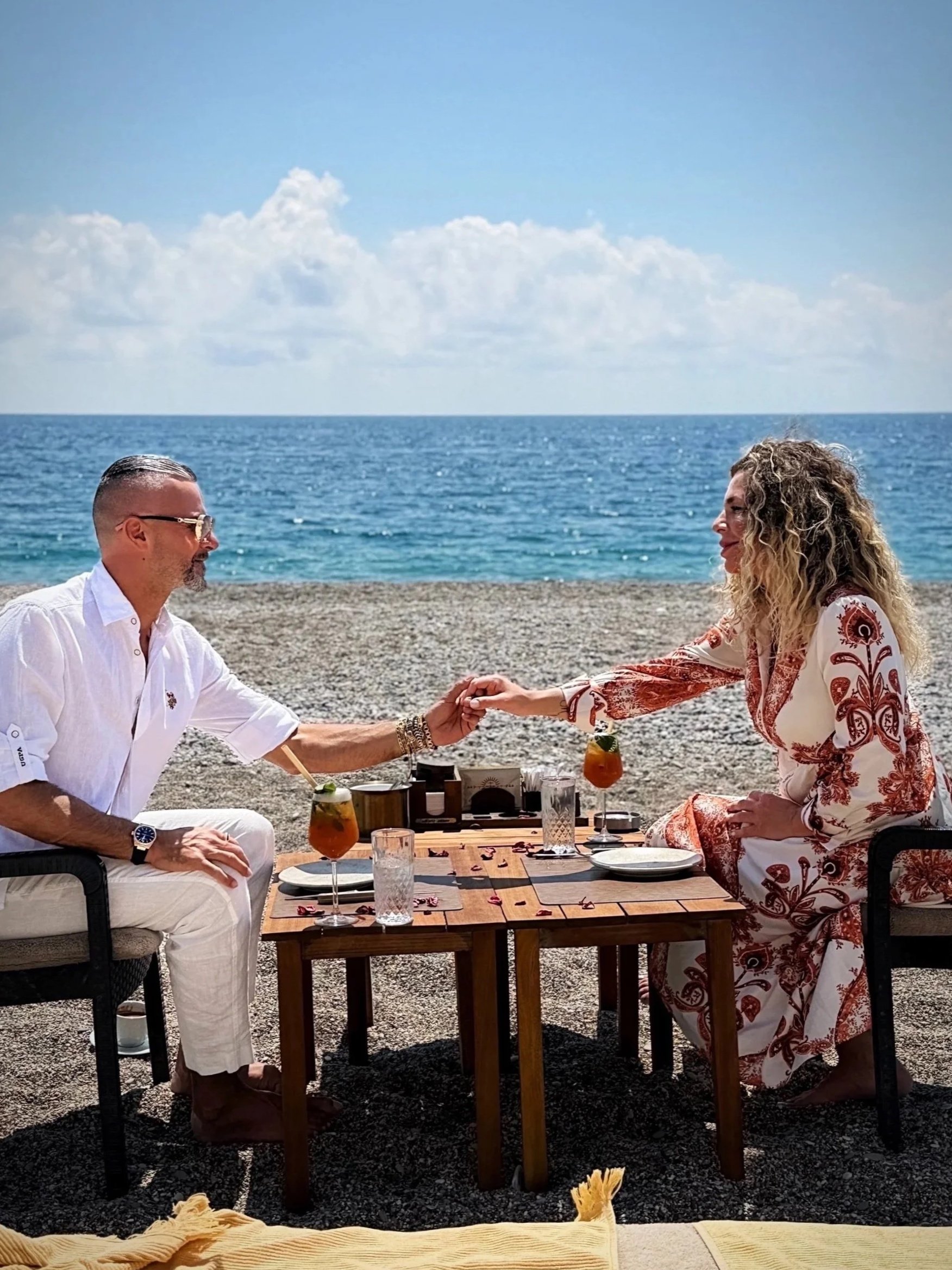 A man and woman sitting at a table on a beach, holding hands and smiling at each other, with the ocean and sky in the background.