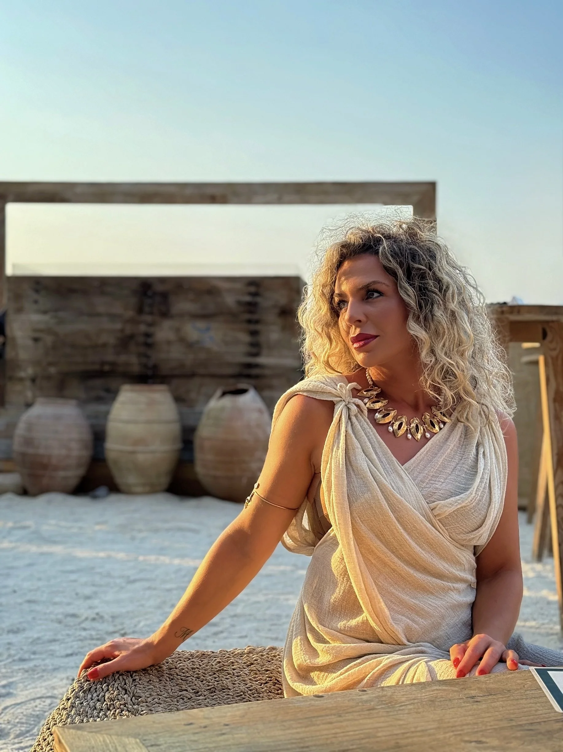 A woman with blonde curly hair, wearing a beige wrap dress and a statement necklace, seated outdoors near water with large decorative vases in the background, during sunset or late afternoon.