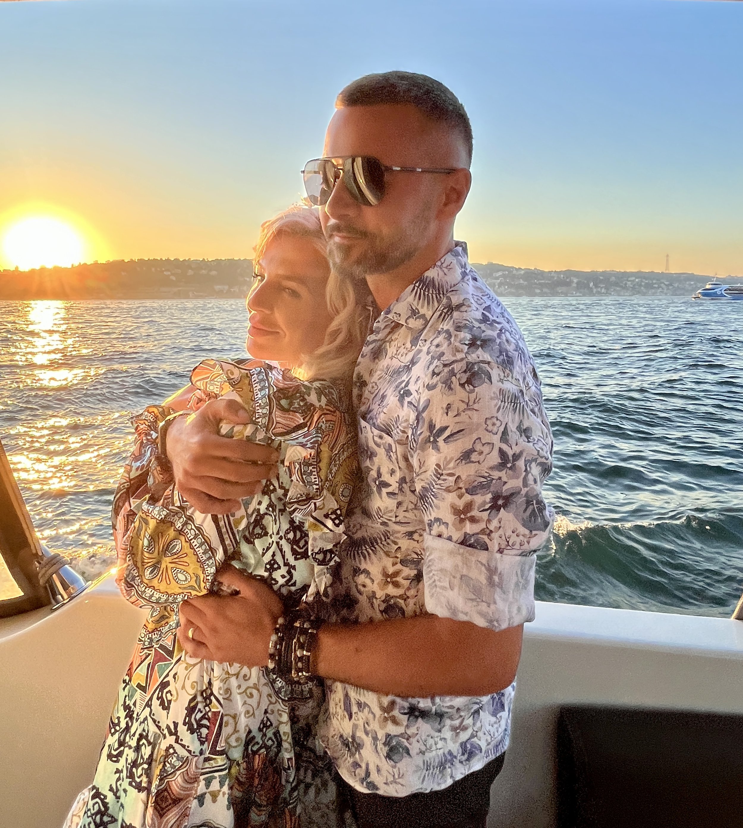 Two people embrace on a boat during sunset, with water and a distant shoreline with buildings and a lighthouse in the background.