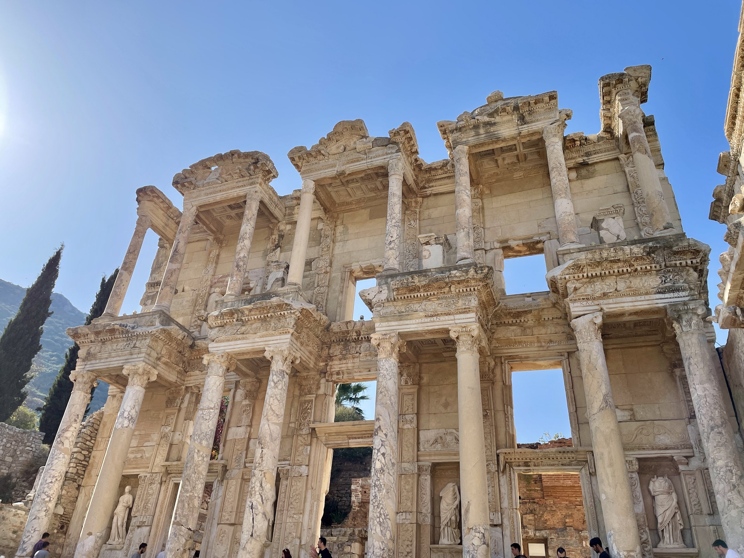 Ancient Greco-Roman ruins with tall columns and detailed carvings under a clear blue sky, with people walking nearby.