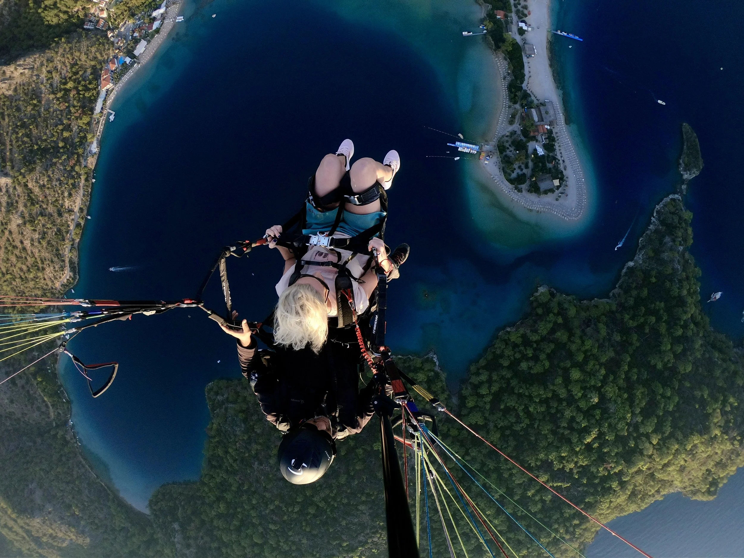 A person paragliding high above a blue water body with small boats and a shoreline with buildings and green trees.