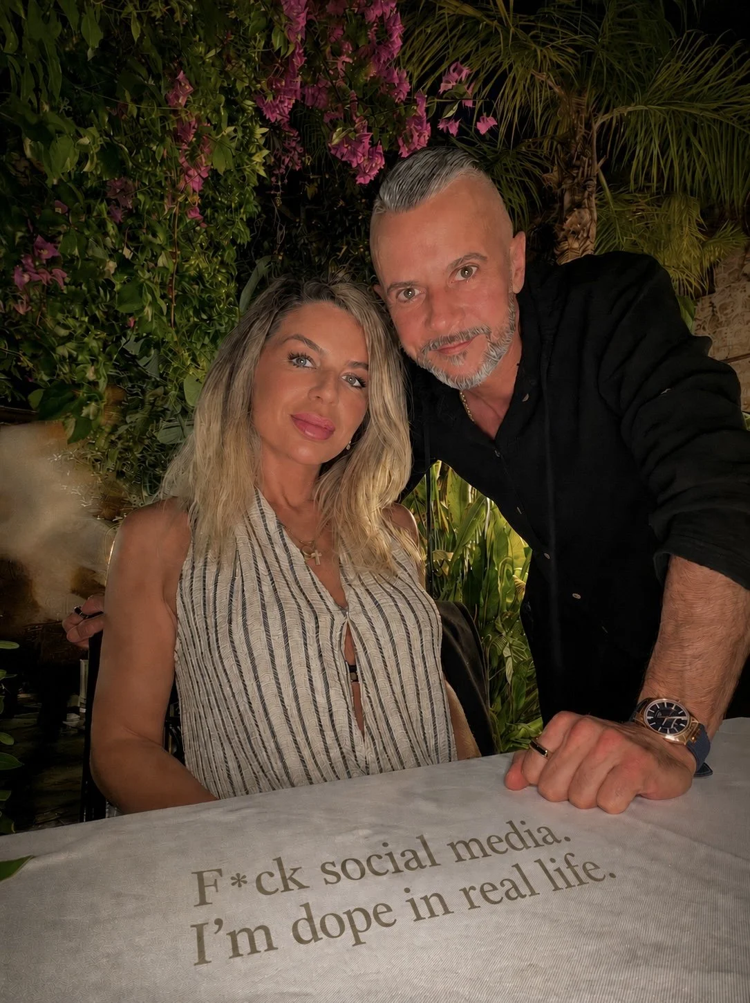 A man and a woman posing together outdoors at night surrounded by lush green plants. The woman is seated, wearing a sleeveless striped top, with wavy blonde hair, and makeup. The man is standing next to her, leaning over with a smile, with short, slicked-back gray hair, a beard, and wearing a black shirt and a watch. A table in front of them has a white cloth with the humorous phrase, 'F*ck social media. I'm dope in real life.'