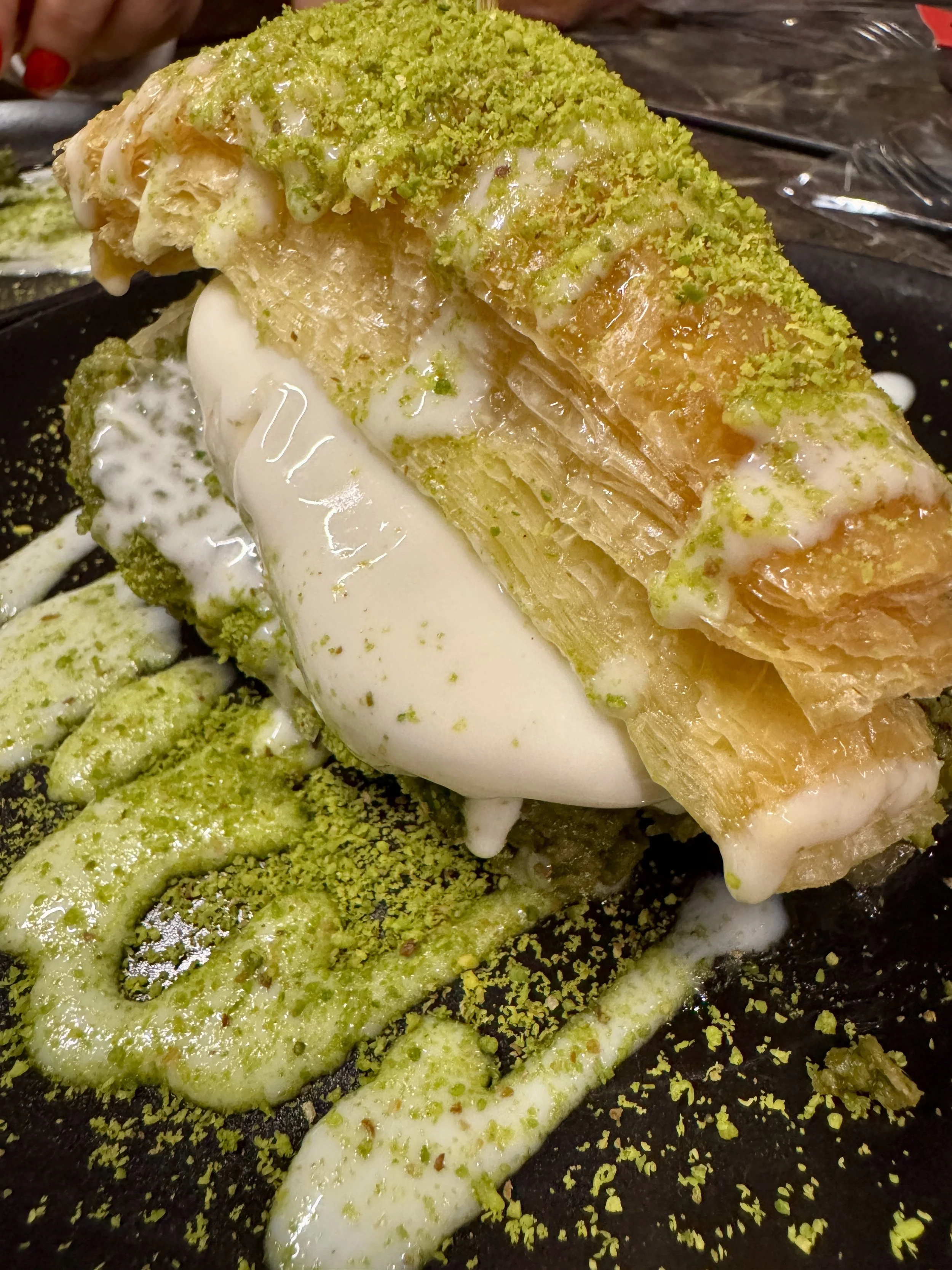A slice of traditional Turkish baklava topped with crushed pistachios, with a layer of white cream or ice cream underneath, presented on a dark plate.